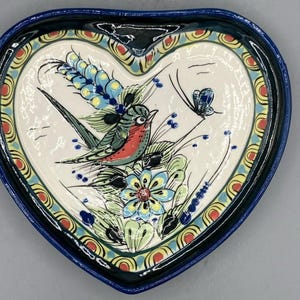 May include: A heart-shaped ceramic dish with a glossy dark blue rim and a cream-colored center. The surface features a hand-painted illustration of a bird with a red chest, a blue butterfly, green foliage, and a large blue flower with yellow petals.