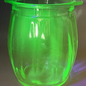 Op de afbeelding: Een doorschijnende, levendige neongroene glazen pot met een tonvormig ontwerp en twee kleine handvatten aan de rand. Het buitenoppervlak is voorzien van een delicaat, geëtst patroon van druiventrossen en dunne ranken rond het midden.