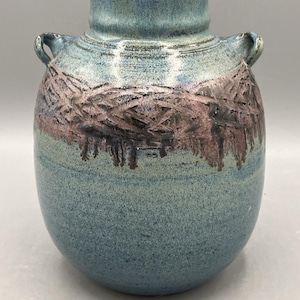 May include: A ceramic vase with a rounded body and two small handles at the neck. The surface features a mottled blue glaze with a textured, dark brown and mauve band encircling the upper section, creating a distinct contrast in color and finish.