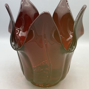 May include: A deep red glass vase with a wide, undulating rim that mimics the shape of an open flower petal. The body of the vase features vertical, curved indentations that extend from the base to the flared, wavy top edge, creating a smooth, translucent surface throughout the piece.