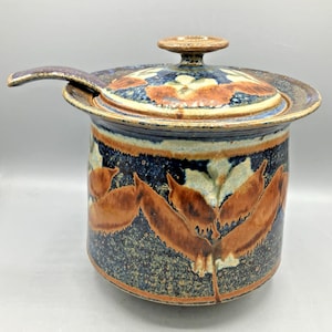 May include: A ceramic lidded jar with a matching ladle featuring a dark blue and brown speckled glaze. The exterior and lid are decorated with a repeating pattern of stylized orange-brown leaves against a deep blue and cream background.