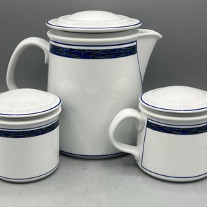 May include: A white ceramic tea set consisting of a large teapot, a lidded sugar bowl, and a lidded creamer. Each piece features a subtle grey diamond pattern on the body and a horizontal band of dark blue with thin gold wavy lines near the top.