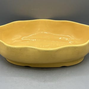 Puede incluir: Un cuenco de cerámica amarillo con forma ovalada y un borde ondulado y festoneado. El cuenco presenta un acabado liso y brillante con un color sólido y uniforme en todas sus superficies interiores y exteriores, sobre una base ovalada.