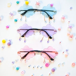 May include: Three pairs of cloud-shaped sunglasses with dangling charms. The sunglasses are in blue, purple, and pink.