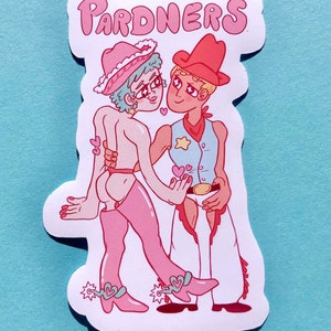 Op de afbeelding: Een sticker met een roze en blauwe illustratie van twee cowboys die elkaars hand vasthouden. De cowboy links draagt een roze hoed en laarzen, en de cowboy rechts draagt een rode hoed en laarzen. De sticker zegt "PARDNERS" in roze letters.