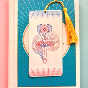 Puede incluir: Un marcapáginas con una ilustración colorida de una figura alada con un ojo en el centro de su cabeza. El marcapáginas tiene un degradado rosa y azul y un borla amarilla.