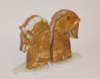 May include: A pair of bookends made of brown and white marble, shaped like horse heads. The bookends are on white marble bases.