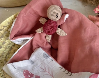 May include: A pink and white muslin baby blanket with a crocheted turtle toy. The blanket has a floral pattern and the turtle is brown with a red shell.