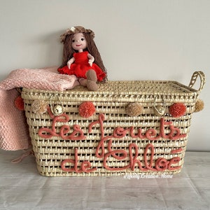 Peut inclure: Un panier de rangement en osier tressé avec une couverture rose et une poupée en crochet posée dessus. Le panier porte l'inscription "Les Jouets de Chloe" en fil rose sur le devant.