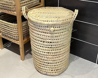 May include: A natural woven laundry hamper with a lid and handles. The hamper is cylindrical and made of light brown woven material. A wooden shelf unit with woven baskets is visible in the background. The hamper is designed for laundry storage.