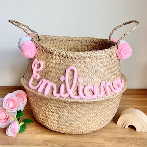 Puede incluir: Una cesta de mimbre tejida con dos asas, decorada con pompones de lana rosa y blanca en los laterales. El nombre Emiliana está escrito en la parte frontal con letras grandes de lana rosa, creando un aspecto texturizado y personalizado.