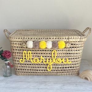 Op de afbeelding: Een rechthoekige opbergmand van gevlochten stro met een deksel en twee handvatten aan de zijkanten. De voorzijde toont de naam Marylou in gele garenletters. Een decoratieve slinger van witte, gele en beige pompons langs de bovenrand.