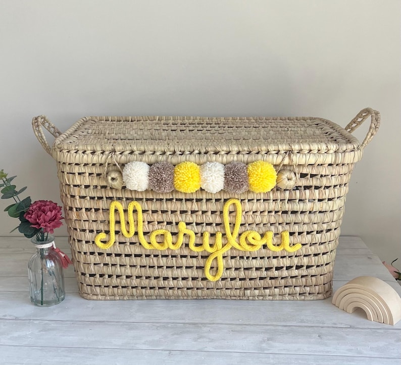 Op de afbeelding: Een rechthoekige opbergmand van gevlochten stro met een deksel en twee handvatten aan de zijkanten. De voorzijde toont de naam Marylou in gele garenletters. Een decoratieve slinger van witte, gele en beige pompons langs de bovenrand.