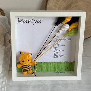 May include: A white frame with a crocheted bee holding balloons. The frame includes the name Mariya, the date 06 juin 2023, the time 08h03, the length 50 cm, and the weight 3kg420. The bottom of the frame has a green grass-like material and the text LovelyCreation.