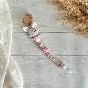 May include: A white crocheted bunny with pink accents and a wooden bunny attached to a clip. The bunny is part of a teething toy with wooden beads, pink and white beads, and wooden letter beads that spell out the name "MANON".