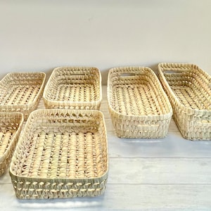 Puede incluir: Seis cestas rectangulares tejidas de materiales naturales. Las cestas son todas del mismo tama&ntilde;o y forma, y est&aacute;n dispuestas en fila sobre una superficie blanca.