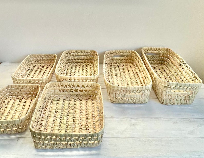 Puede incluir: Seis cestas rectangulares tejidas de materiales naturales. Las cestas son todas del mismo tama&ntilde;o y forma, y est&aacute;n dispuestas en fila sobre una superficie blanca.
