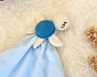 May include: A baby security blanket featuring a crocheted turtle with a blue shell and cream-colored flippers and head. The blanket is a soft, light blue color.