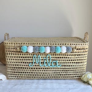 Op de afbeelding: Een rechthoekige opbergmand van gevlochten stro met een deksel en twee handvatten aan de zijkanten. De voorzijde heeft een rij afwisselende turquoise, witte en grijze pompons boven de naam Mila in turquoise cursief schrift.