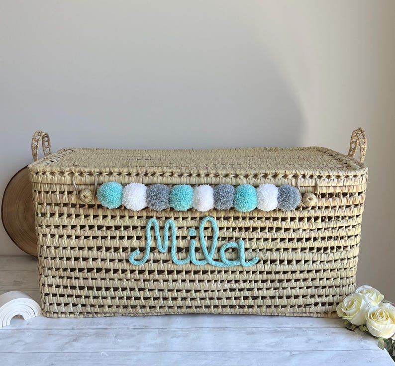 Op de afbeelding: Een rechthoekige opbergmand van gevlochten stro met een deksel en twee handvatten aan de zijkanten. De voorzijde heeft een rij afwisselende turquoise, witte en grijze pompons boven de naam Mila in turquoise cursief schrift.