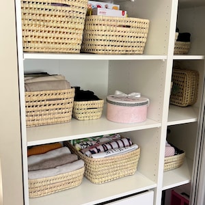 Könnte beinhalten: Weiße Schrankregale mit geflochtenen Körben, gefüllt mit gefalteten Kleidungsstücken und einer rosafarbenen Geschenkbox. Die Regale sind ordentlich und aufgeräumt.