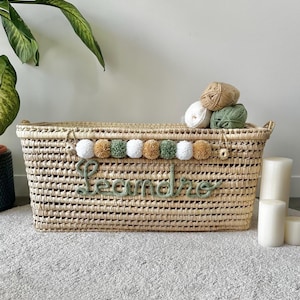 Op de afbeelding: Een rechthoekige mand van gevlochten stro met een decoratieve rij witte, beige en saliegroene pompons. De naam Leandro is in saliegroene cursieve garenletters op de voorzijde aangebracht. Drie bollen wol liggen bovenop het deksel van de mand.