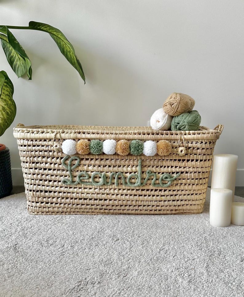 Op de afbeelding: Een rechthoekige mand van gevlochten stro met een decoratieve rij witte, beige en saliegroene pompons. De naam Leandro is in saliegroene cursieve garenletters op de voorzijde aangebracht. Drie bollen wol liggen bovenop het deksel van de mand.