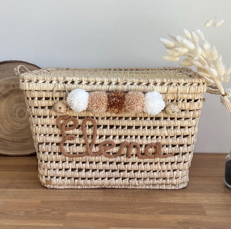 Op de afbeelding: Een rechthoekige mand van gevlochten stro met een deksel en een rij van vijf decoratieve pompons in wit en bruintinten. De naam Elena is in bruine, cursieve garenletters op de voorzijde van de natuurkleurige mand aangebracht.