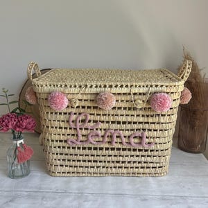 Peut inclure: Un panier de rangement en osier naturel avec une bordure de pompons roses et le nom "Lena" brodé en fil rose.