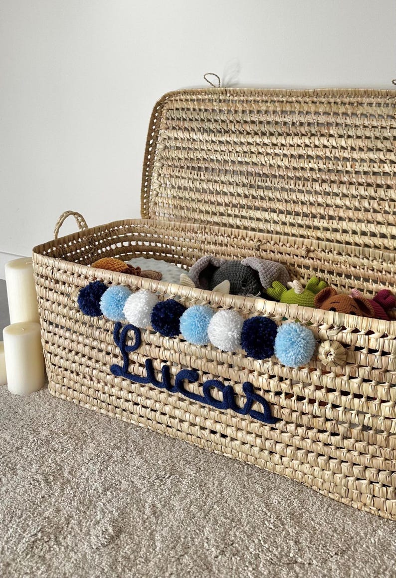 Op de afbeelding: Een rechthoekige opbergmand van gevlochten stro met een scharnierend deksel, voorzien van een rij afwisselend marineblauwe, lichtblauwe en witte pompons. De naam Lucas is in donkerblauw cursief schrift op de voorzijde geborduurd.