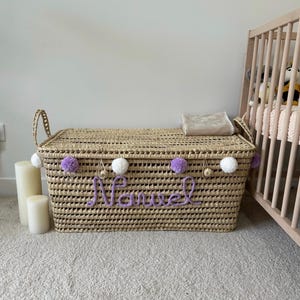 Puede incluir: Una cesta de almacenamiento rectangular tejida con tapa y dos asas laterales. El frente está decorado con pompones blancos y lavanda alternados y el nombre "Marvel" escrito en letras cursivas de lana color lavanda en el centro.