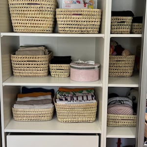 Puede incluir: Un armario blanco con varios estantes y cajones. Los estantes est&aacute;n llenos de cestas tejidas, algunas con ropa doblada y otras con juguetes. Los cajones est&aacute;n vac&iacute;os. El armario est&aacute; abierto y el interior es visible.