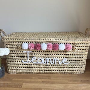Op de afbeelding: Een rechthoekige opbergmand van gevlochten stro met een deksel en twee handvatten aan de zijkanten. De voorzijde is voorzien van een rij afwisselend witte en roze pompons en de naam Jeanne in witte cursieve letters.