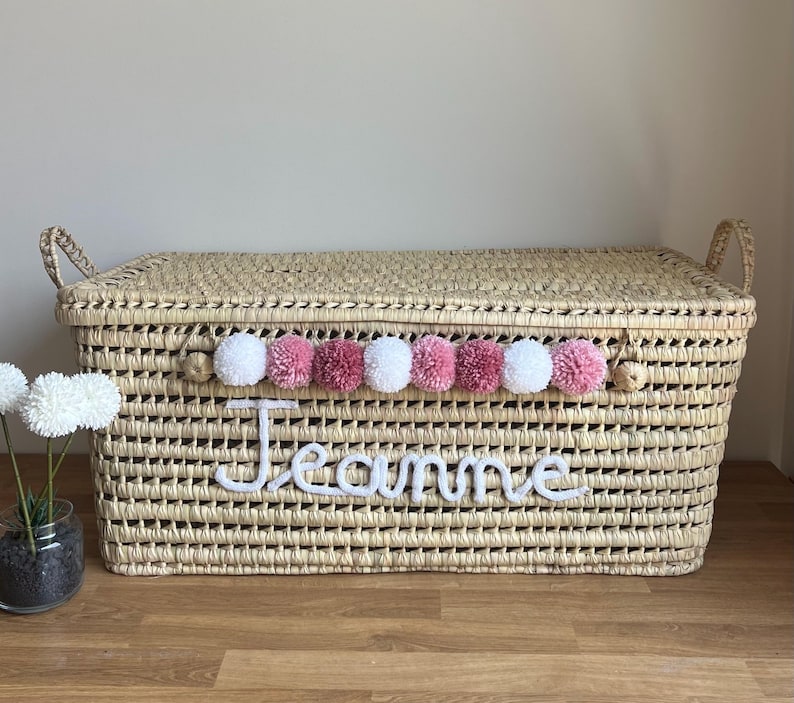 Op de afbeelding: Een rechthoekige opbergmand van gevlochten stro met een deksel en twee handvatten aan de zijkanten. De voorzijde is voorzien van een rij afwisselend witte en roze pompons en de naam Jeanne in witte cursieve letters.
