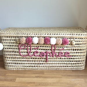 Op de afbeelding: Een rechthoekige opbergmand van gevlochten stro met een deksel en twee handvatten aan de zijkanten. De voorzijde is voorzien van een rij afwisselende beige, roze en witte pompons boven de naam Cl&eacute;oph&eacute;e in roze gebreide cursieve letters.
