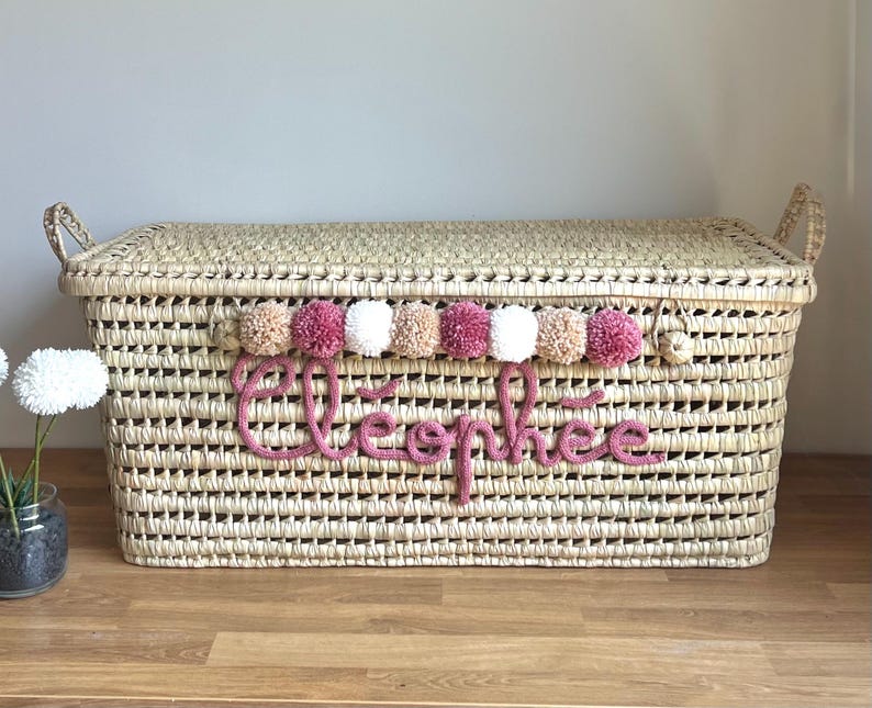 Op de afbeelding: Een rechthoekige opbergmand van gevlochten stro met een deksel en twee handvatten aan de zijkanten. De voorzijde is voorzien van een rij afwisselende beige, roze en witte pompons boven de naam Cl&eacute;oph&eacute;e in roze gebreide cursieve letters.