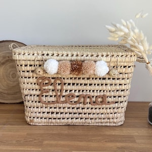 Puede incluir: Una cesta rectangular de paja trenzada con tapa y una fila de cinco pompones alternos en blanco y beige. El nombre Elena está escrito en letras cursivas de lana marrón en la parte frontal de la cesta de color natural.