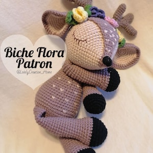 Peut inclure: Un jouet en crochet en forme de cerf avec un corps violet, des sabots noirs et une couronne de fleurs. Le cerf est couché sur le côté, les yeux fermés. Le texte "Biche Flora Patron @LovelyCreation_Home" est écrit sur l'image.