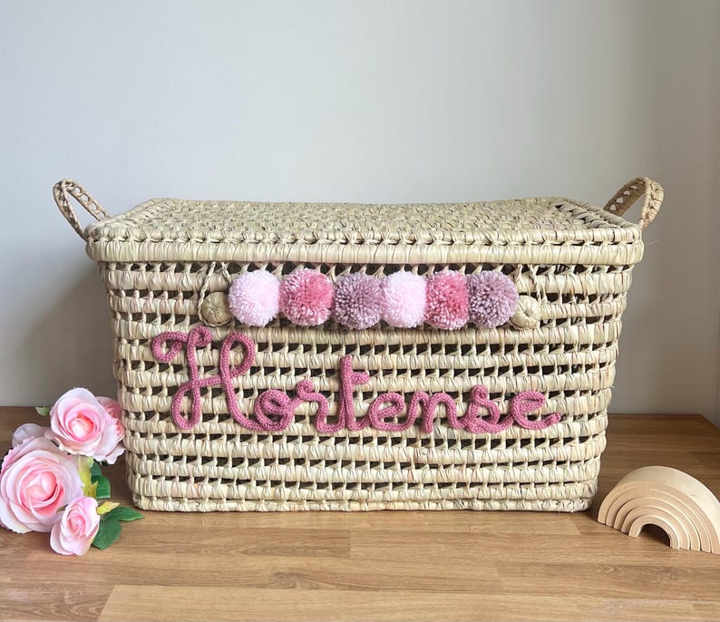 Op de afbeelding: Een rechthoekige mand van gevlochten stro met een deksel en twee handvatten aan de zijkanten. De voorzijde is voorzien van een rij van zes roze en mauve wollen pompons en de naam Hortense in cursieve roze wollen letters.