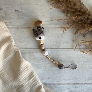 Può includere: Una testa di volpe in crochet grigio e bianco attaccata a un mordedor per bambini in legno con un perno di legno, un perno di crochet bianco e un perno di legno con il nome "LOUP" scritto su di esso.