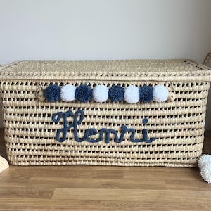 Op de afbeelding: Een rechthoekige opbergmand van gevlochten stro met een deksel en twee handvatten aan de zijkanten. De voorzijde is voorzien van een rij afwisselende marineblauwe en witte wollen pompons en de naam Henri in marineblauw cursief garen.