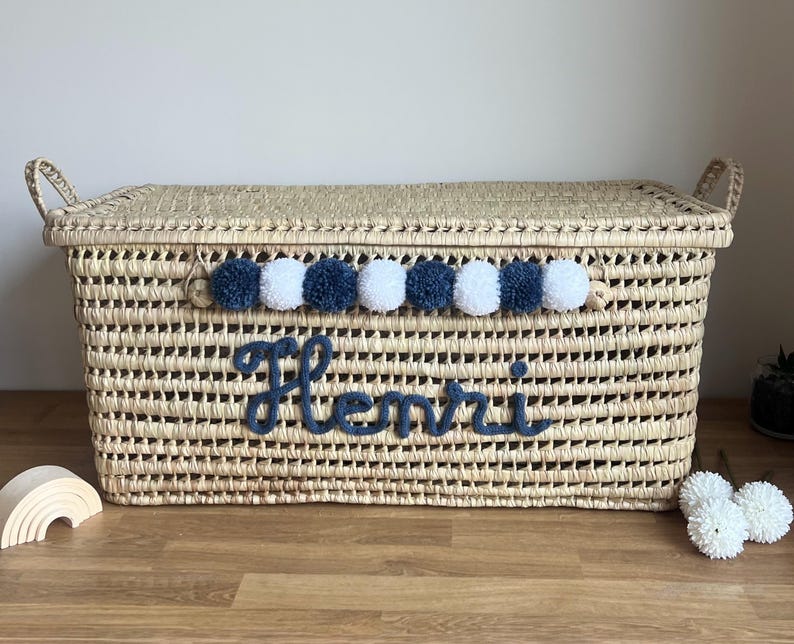 Op de afbeelding: Een rechthoekige opbergmand van gevlochten stro met een deksel en twee handvatten aan de zijkanten. De voorzijde is voorzien van een rij afwisselende marineblauwe en witte wollen pompons en de naam Henri in marineblauw cursief garen.