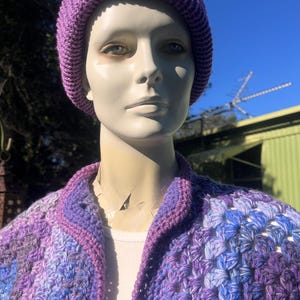 May include: A purple knit beanie and a crocheted cardigan in shades of purple and blue are displayed on a mannequin. The cardigan has a button closure and a contrasting purple trim. The beanie is a solid, ribbed knit.