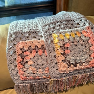 May include: A handmade crochet blanket in a granny square pattern. The blanket features squares in shades of gray, pink, yellow, and brown. It has a fringe along the bottom edge. The blanket is folded and resting on a tan surface.