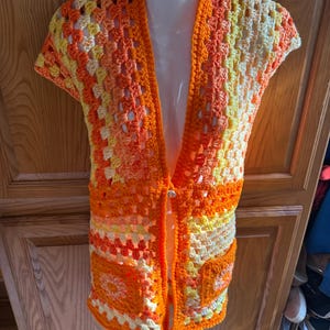 May include: A handmade, open-front crochet vest in shades of orange, yellow, and cream. The vest features a granny square design with short sleeves and two front pockets. The garment is displayed on a white mannequin.