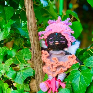 Puede incluir: Una mu&ntilde;eca de crochet con hilo negro para la cara y el cuerpo, hilo rosa para el pelo y un vestido de color melocot&oacute;n con volantes. La mu&ntilde;eca lleva zapatos rosas y est&aacute; sentada en un fondo verde frondoso.