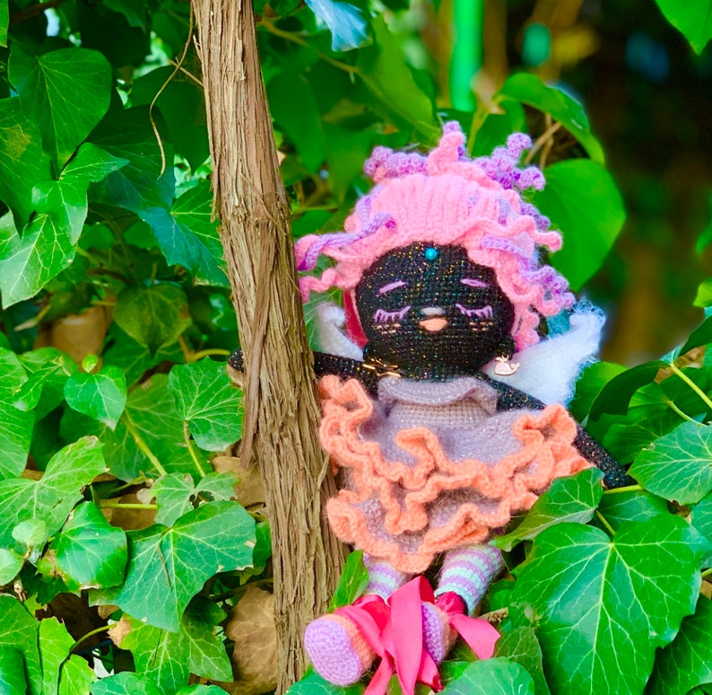 Puede incluir: Una mu&ntilde;eca de crochet con hilo negro para la cara y el cuerpo, hilo rosa para el pelo y un vestido de color melocot&oacute;n con volantes. La mu&ntilde;eca lleva zapatos rosas y est&aacute; sentada en un fondo verde frondoso.
