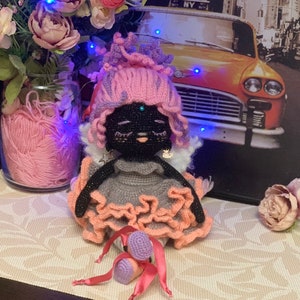 Puede incluir: Una mu&ntilde;eca de crochet con pelo rosa, un vestido gris y zapatos rosa y morado. La mu&ntilde;eca tiene una cara negra con una gema azul en la frente.