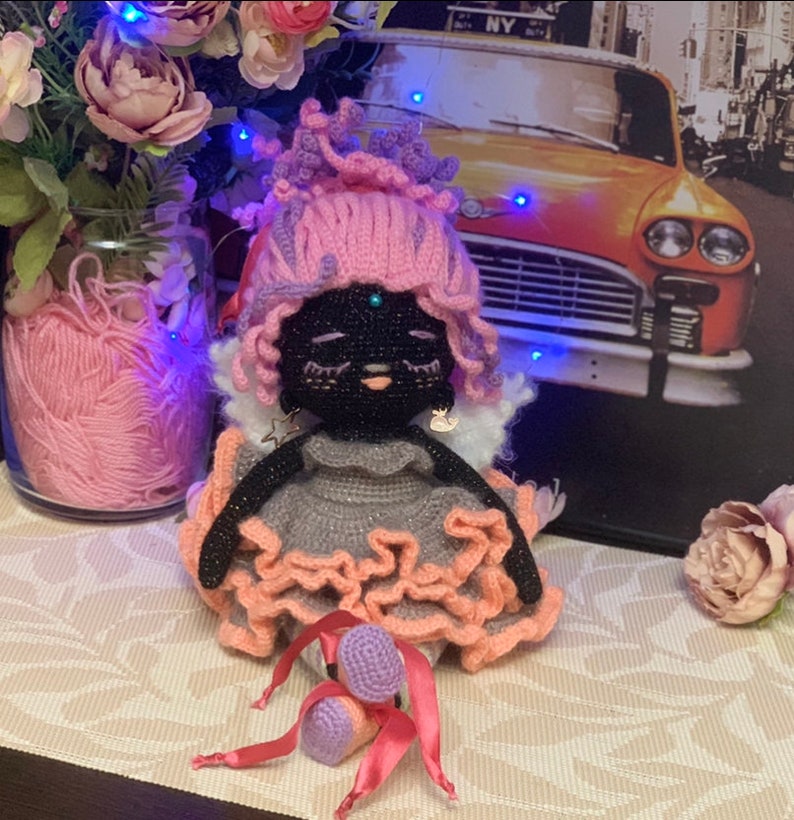 Puede incluir: Una mu&ntilde;eca de crochet con pelo rosa, un vestido gris y zapatos rosa y morado. La mu&ntilde;eca tiene una cara negra con una gema azul en la frente.