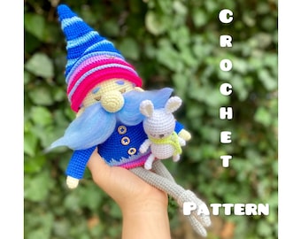 May include: Crochet pattern for a blue and pink gnome holding a white bunny. The gnome has a blue hat with white stripes, a pink and white striped hat, a blue beard, and a blue jacket with white buttons. The bunny has a green scarf.